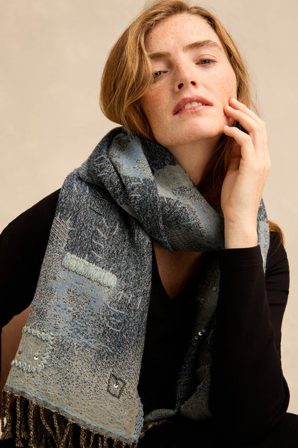 A woman in a black top wears the Etched Patchwork Sequin Wool Scarf—a textured blue wool scarf featuring stitched details, subtle patchwork design, and fringe—as she poses with her hand gently touching her face.