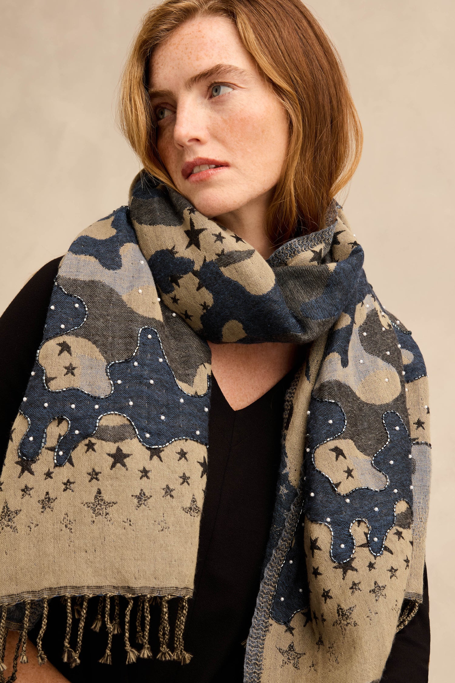 A woman with light brown hair wears the Night Sky Sequin Wool Scarf, featuring a celestial motif in blue and beige tones, draped over a black top as she looks to the side with a neutral expression.