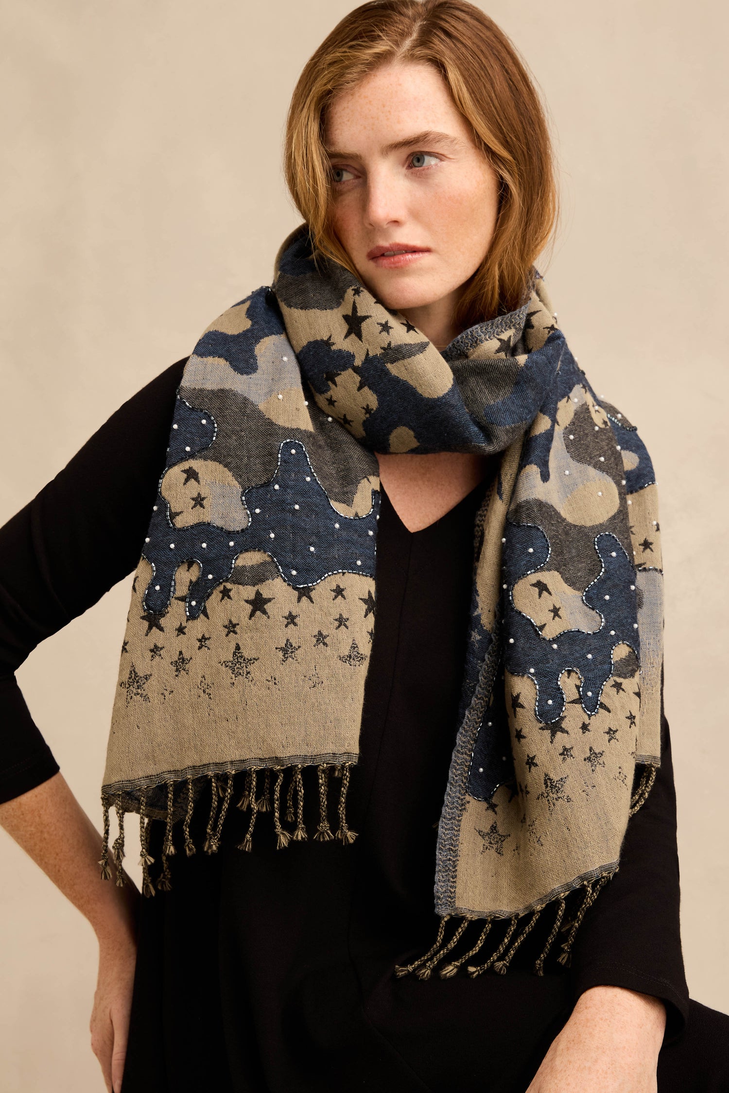 A woman wearing the Night Sky Sequin Wool Scarf—a large, patterned wool scarf with a celestial motif of stars and subtle camouflage accents in blue, gray, and tan tones—over a black top.