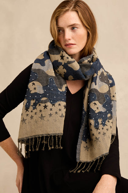A woman wearing the Night Sky Sequin Wool Scarf—a large, patterned wool scarf with a celestial motif of stars and subtle camouflage accents in blue, gray, and tan tones—over a black top.