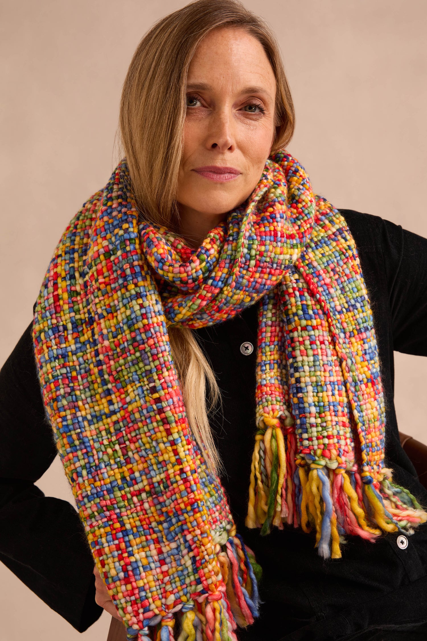 A woman sits facing the camera against a neutral background, wearing the Rainbow Tassel Scarf in a large, multicolored design with fringe. Crafted from soft brushed yarn, this statement accessory layers over her black top, offering lightweight warmth and vibrant flair to any outfit.