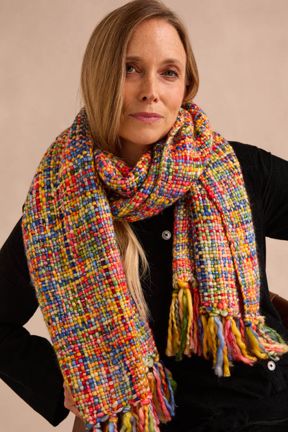 A woman sits facing the camera against a neutral background, wearing the Rainbow Tassel Scarf in a large, multicolored design with fringe. Crafted from soft brushed yarn, this statement accessory layers over her black top, offering lightweight warmth and vibrant flair to any outfit.