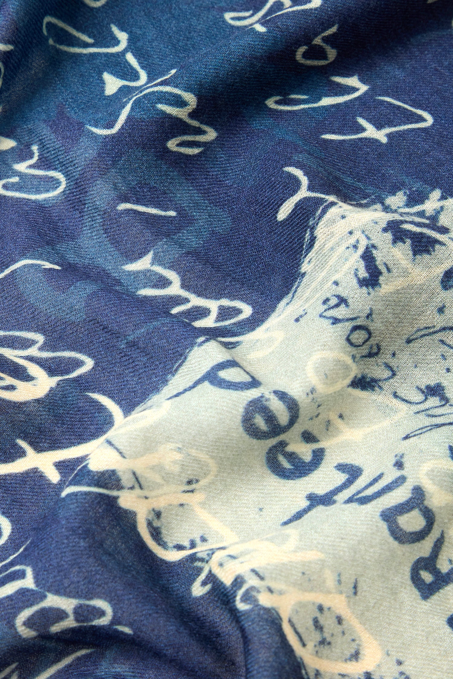 A close-up image of the Love Language Printed Scarf reveals fabric adorned with love letter prints in blue and white hues, featuring cursive script letters scattered randomly across the surface, evoking a vintage-inspired design.