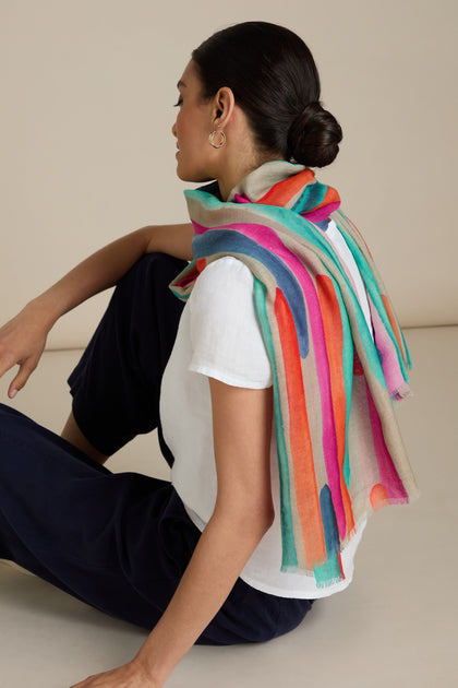 A woman with a bun sits sideways wearing a white top and dark pants. Draped over her shoulders is the Painterly Stripe Wool Mix Scarf, adorned with vibrant brushstroke-inspired stripes, making it perfect for all-occasion styling.