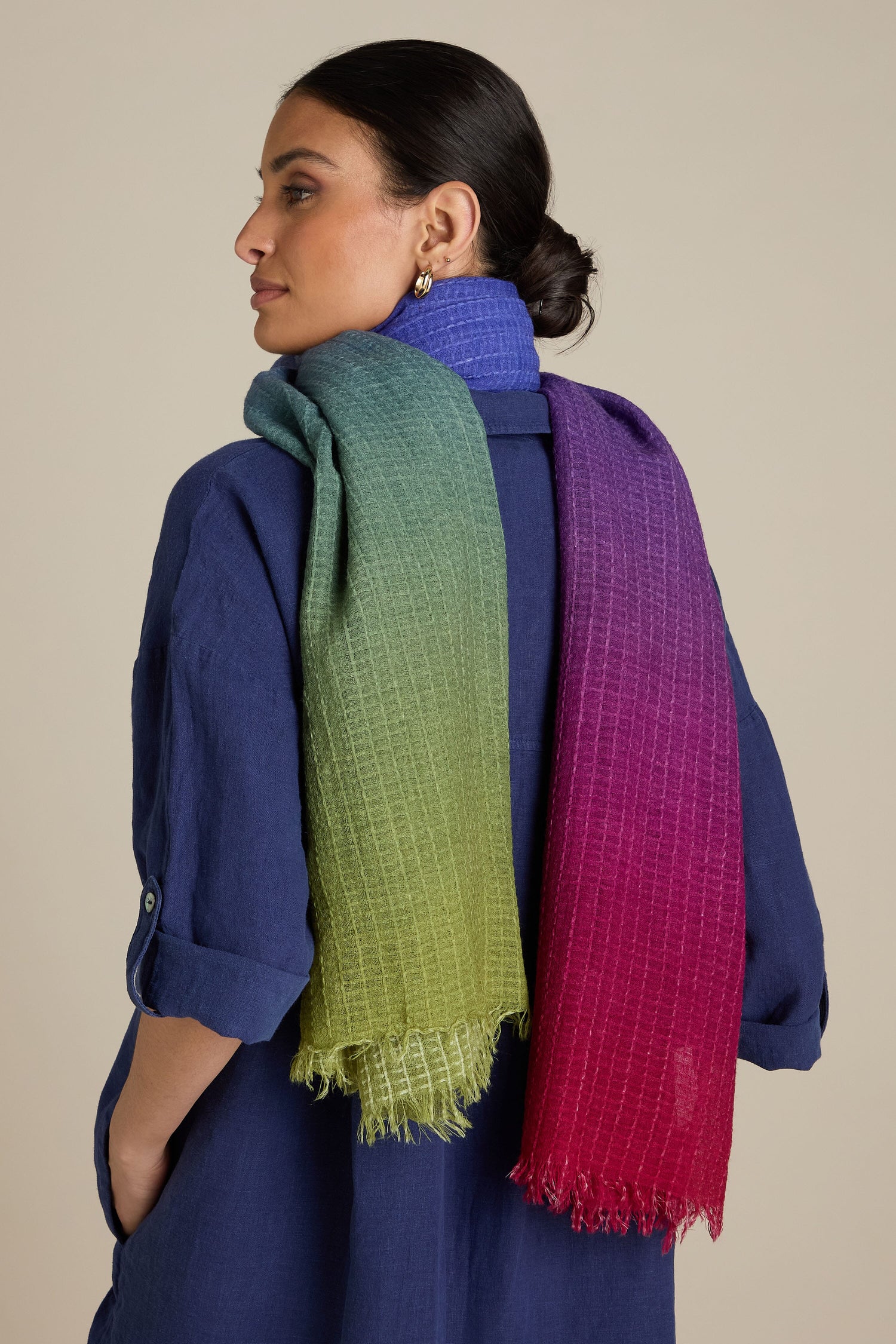 A person with dark hair stands sideways, wearing a blue long-sleeved shirt and a Soft Colour Blend Wool Scarf draped over their shoulder, transitioning in an ombre of jewel tones from green to blue to purple to red.