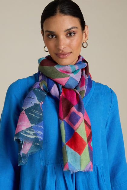 Person wearing a blue top, the Painted Squares Wool Mix Scarf with a colorful geometric motif, and gold hoop earrings, against a neutral background.