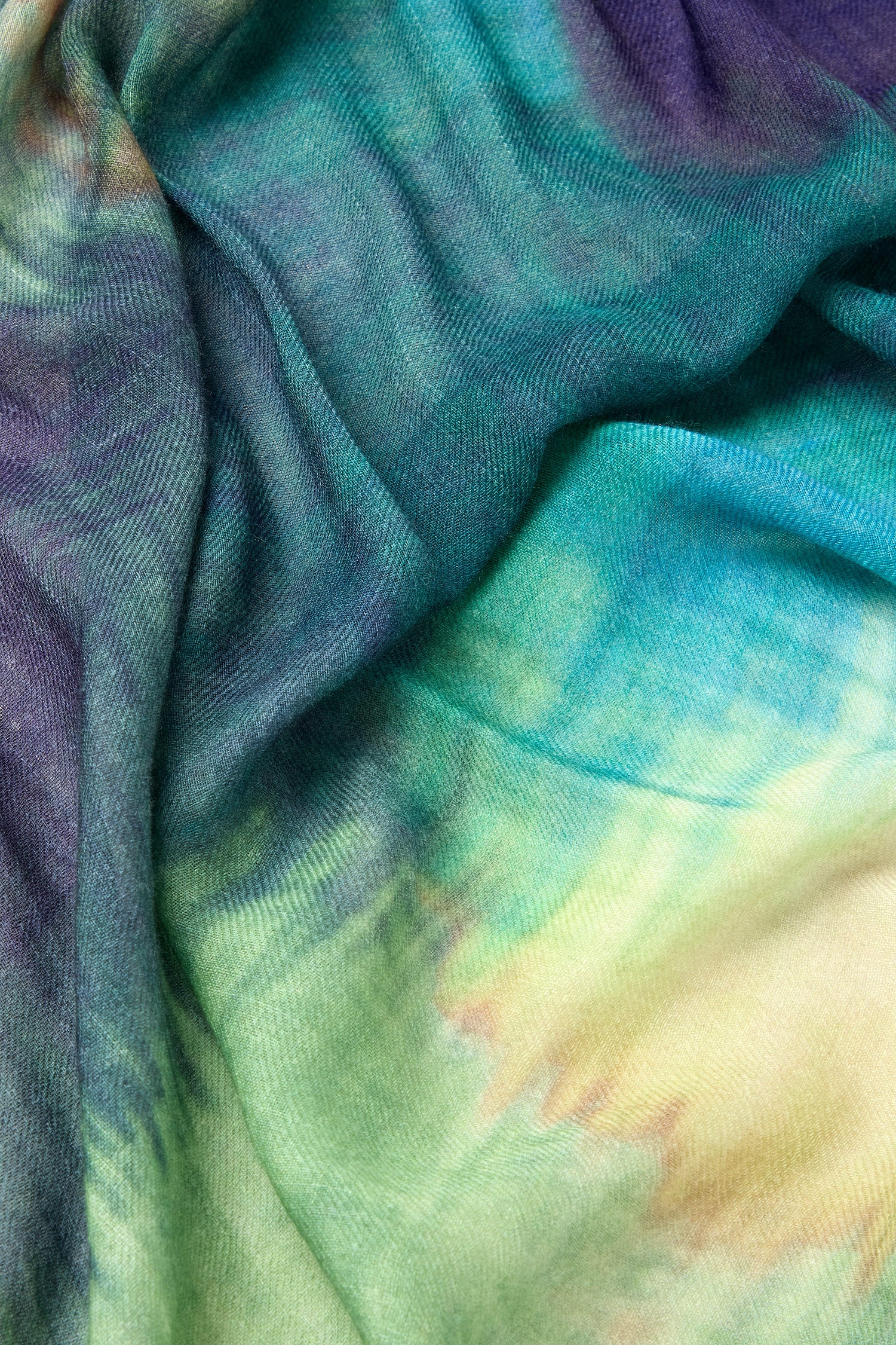 A close-up of the Colour Blend Circle Print Scarf reveals a vibrant tie-dye pattern showcasing shades of green, blue, yellow, and hints of purple. The watercolour-inspired design highlights the sumptuously soft fabric's flowing texture.