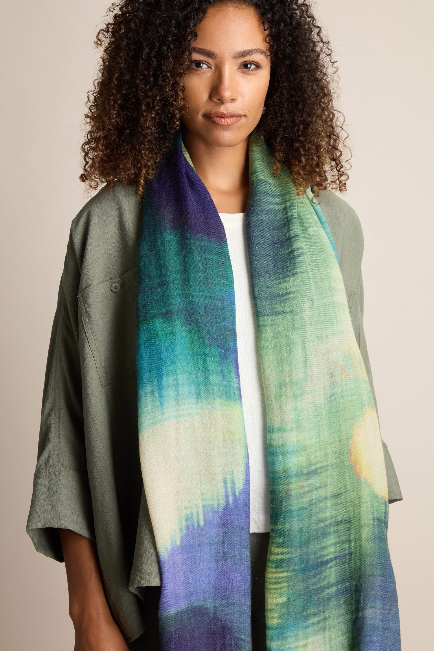 A person with curly hair is wearing a green jacket over a white top and a Colour Blend Circle Print Scarf, which features shades of blue, green, and yellow. The scarf is crafted from sumptuously soft fabric with a watercolor-inspired design. They are standing against a neutral background.