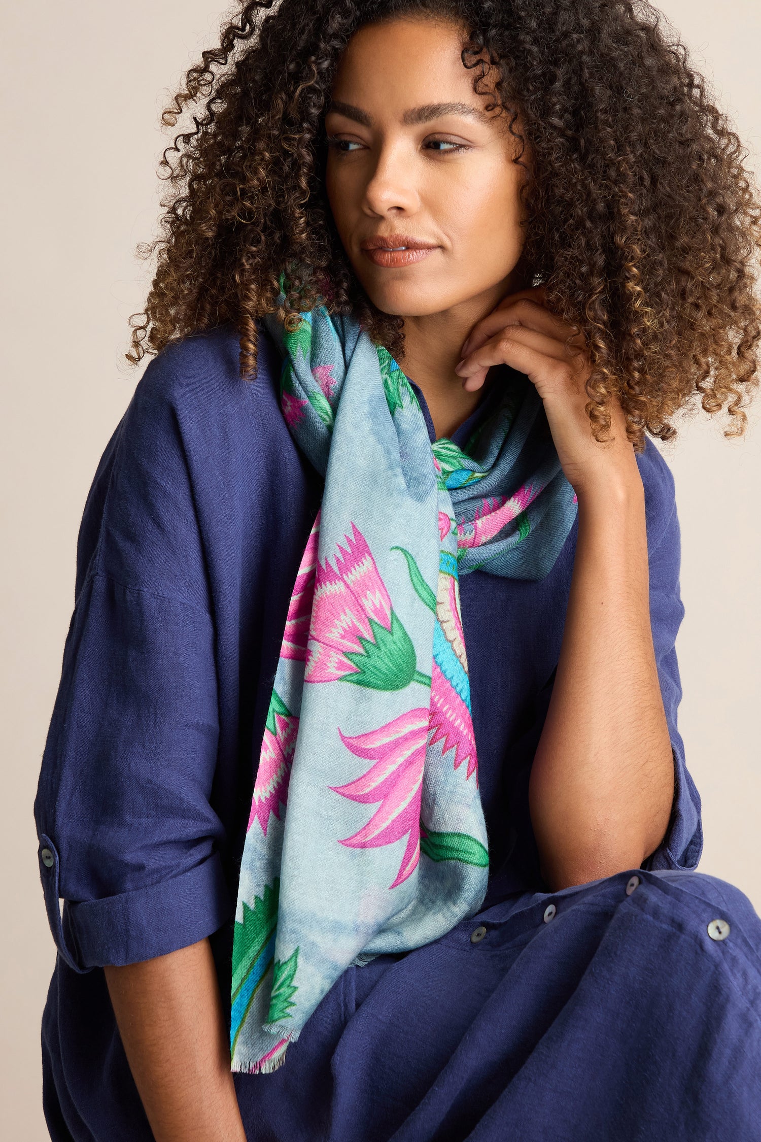 A person with curly hair dons a blue shirt and a lightweight Heavenly Garden Scarf, gazing off to the side.