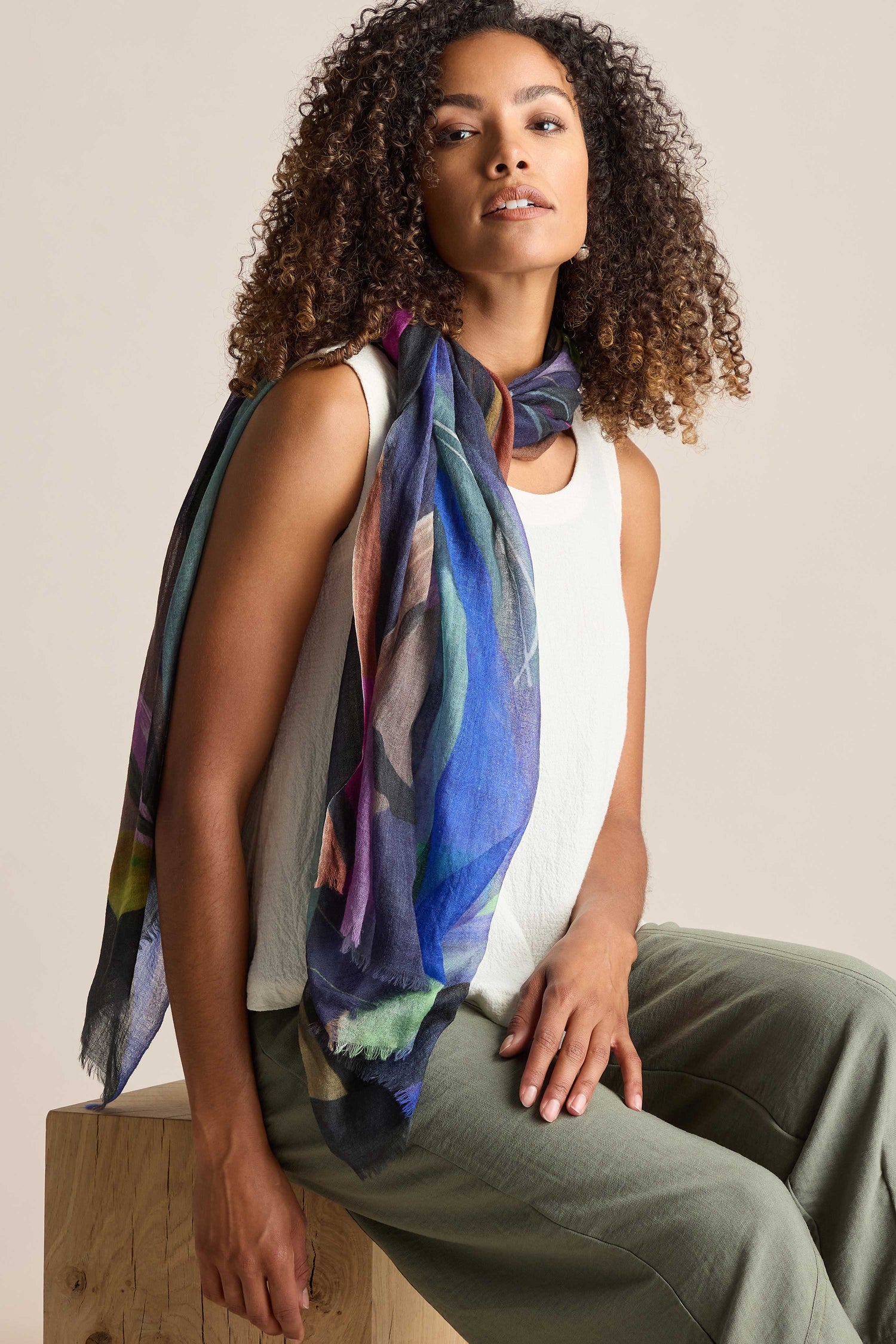 A person with curly hair wearing a colorful, hand-painted Abstract Plume Wool Scarf, a white top, and green pants sits on a wooden block against a plain background.