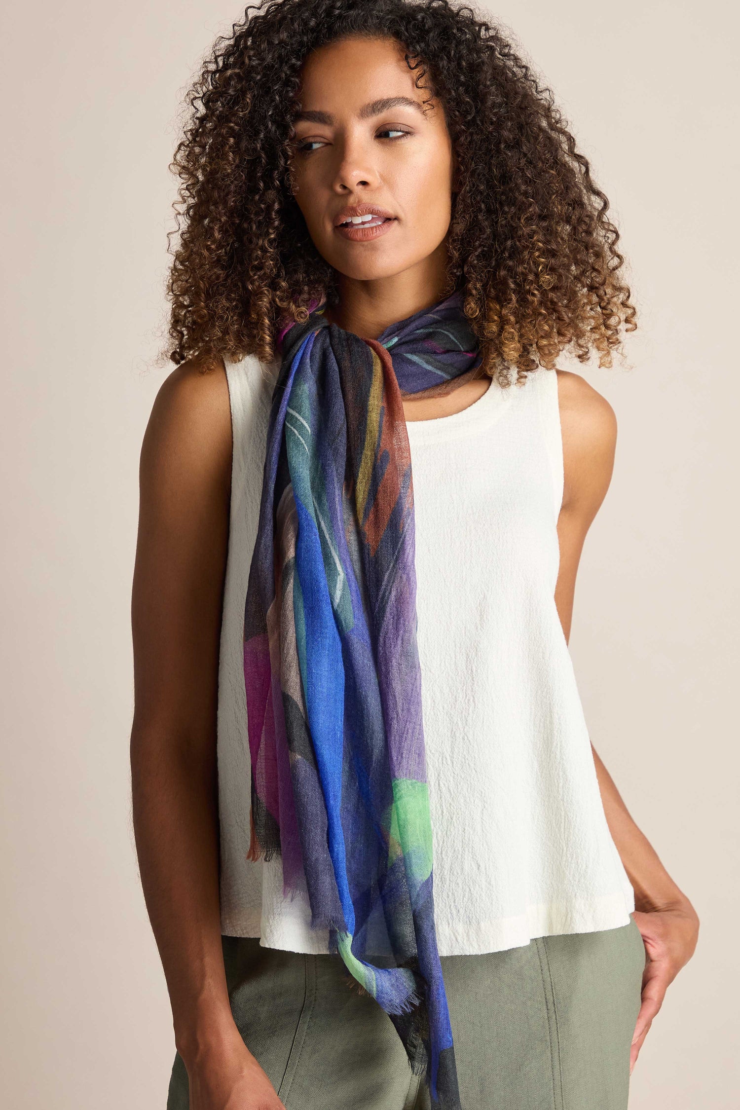 A woman with curly hair wearing a white sleeveless top and the Abstract Plume Wool Scarf, with her left hand in the pocket of her green pants.