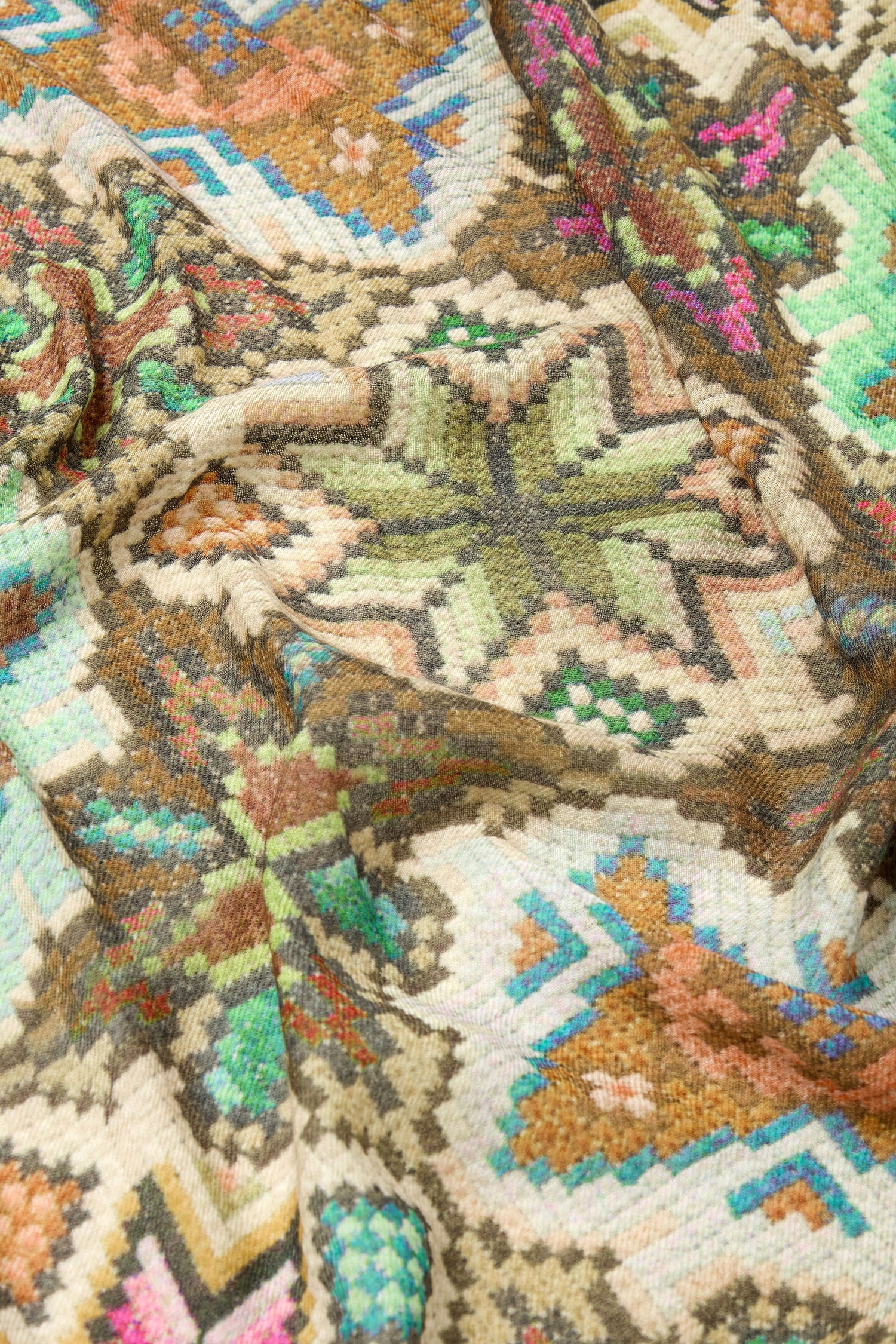 Close-up of a colorful, patterned fabric featuring intricate geometric designs in green, brown, blue, and pink hues. The fabric, reminiscent of a Mosaic Print Wool Mix Scarf, is slightly wrinkled, showing texture and detail.