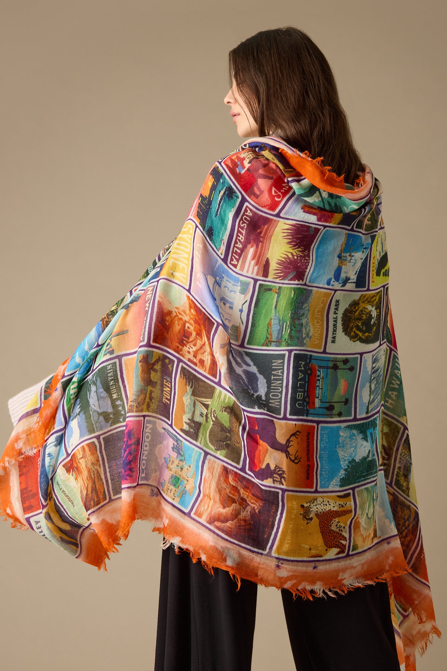 A person draped in the "Postcards From Around the World" wool scarf, showcasing a vibrant collection of travel-themed images, stands against a plain background, exuding an undeniable sense of wanderlust.