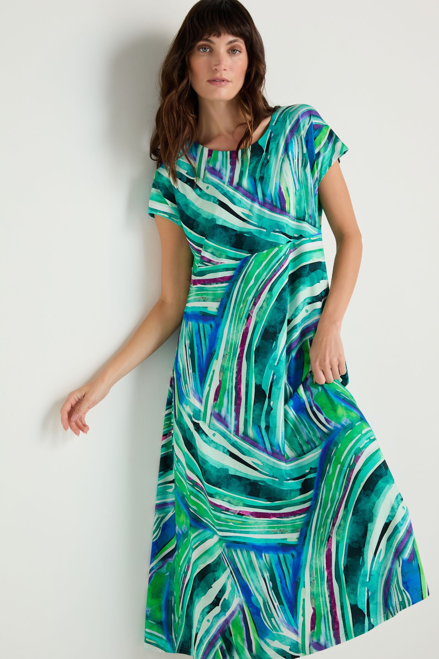 A woman stands against a white wall wearing the Blurred Lines Swing Dress, made from stretch jersey and featuring a green, blue, and white abstract pattern.