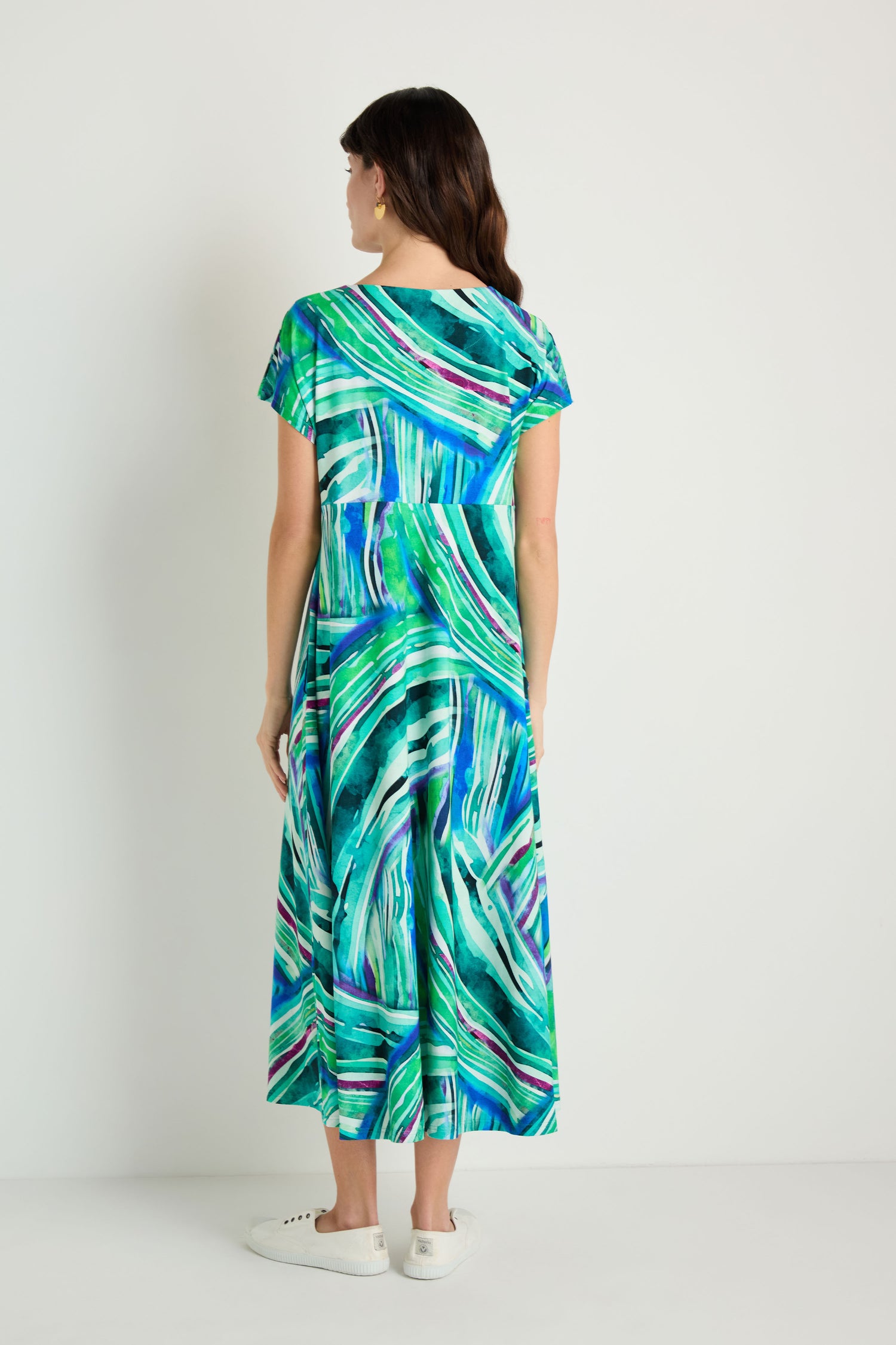 A woman stands against a plain background, facing away, wearing the Blurred Lines Swing Dress—a green and blue abstract patterned stretch jersey midi dress—paired with white shoes.