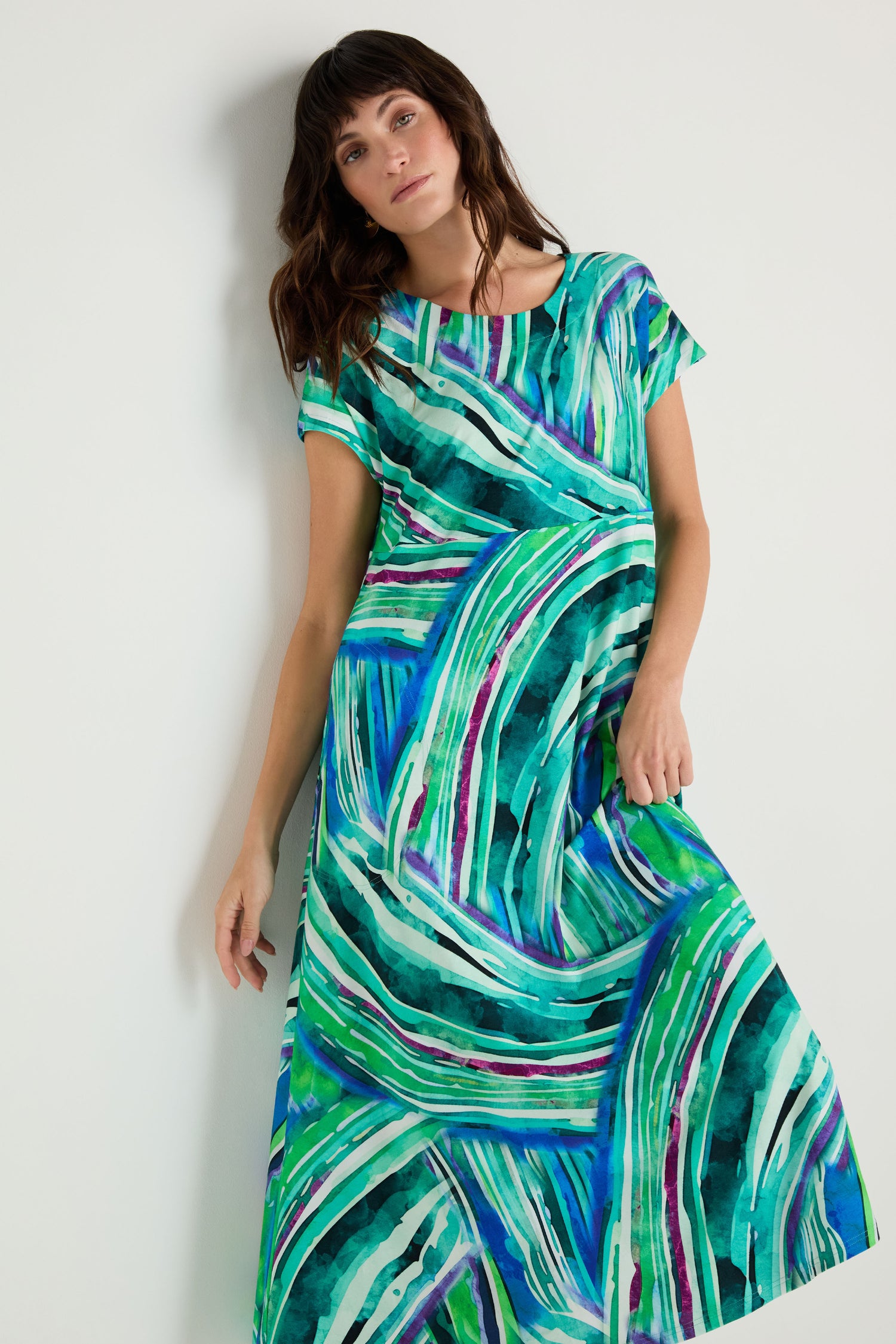 A woman models the Blurred Lines Swing Dress, a loose-fit jersey piece showcasing an abstract green, blue, and white pattern, set against a plain light background.