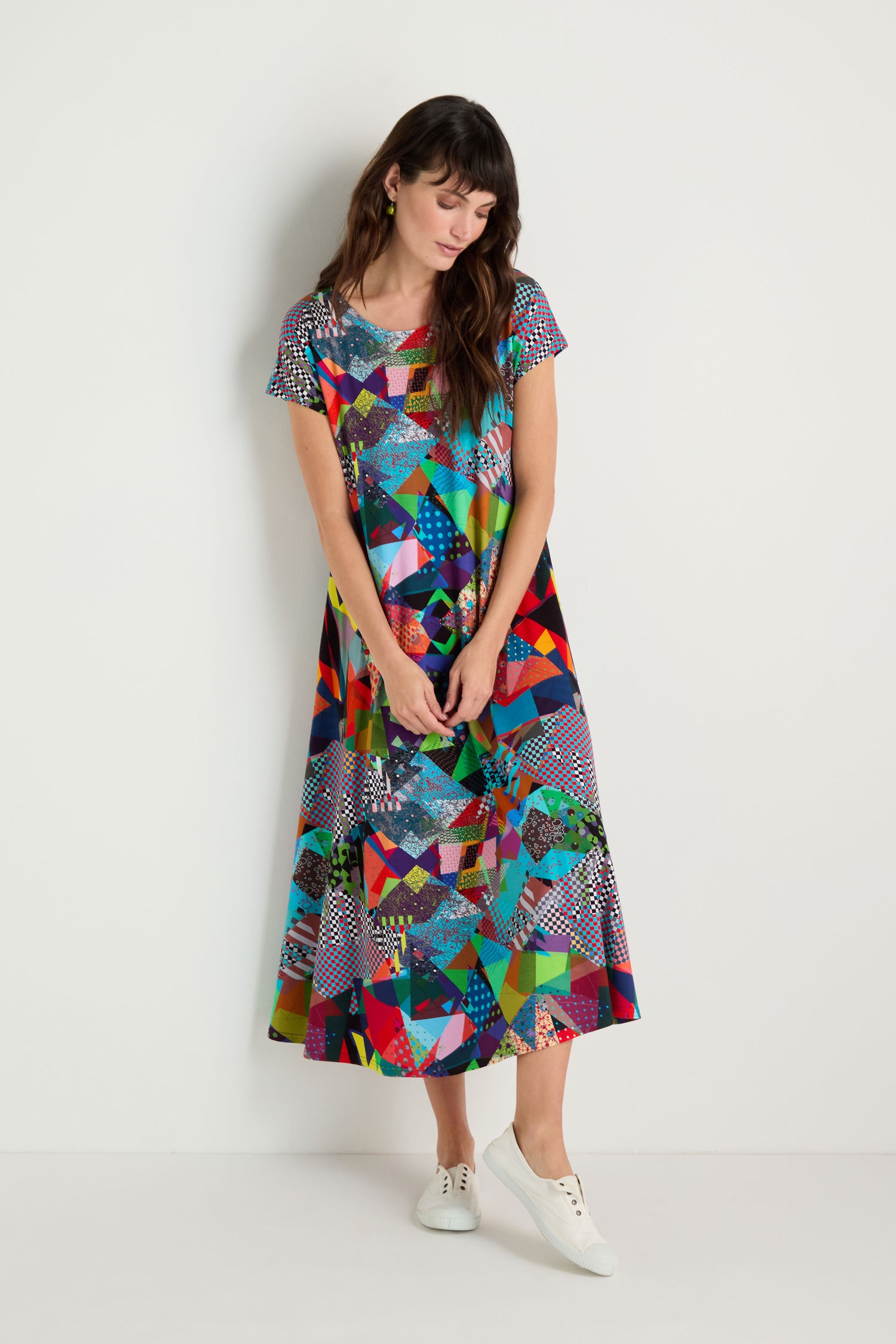 A woman stands against a white wall wearing the Quilt Print Flared Dress—featuring colorful geometric patterns, short sleeves, and paired with white sneakers. She looks down with hands loosely clasped in front.
