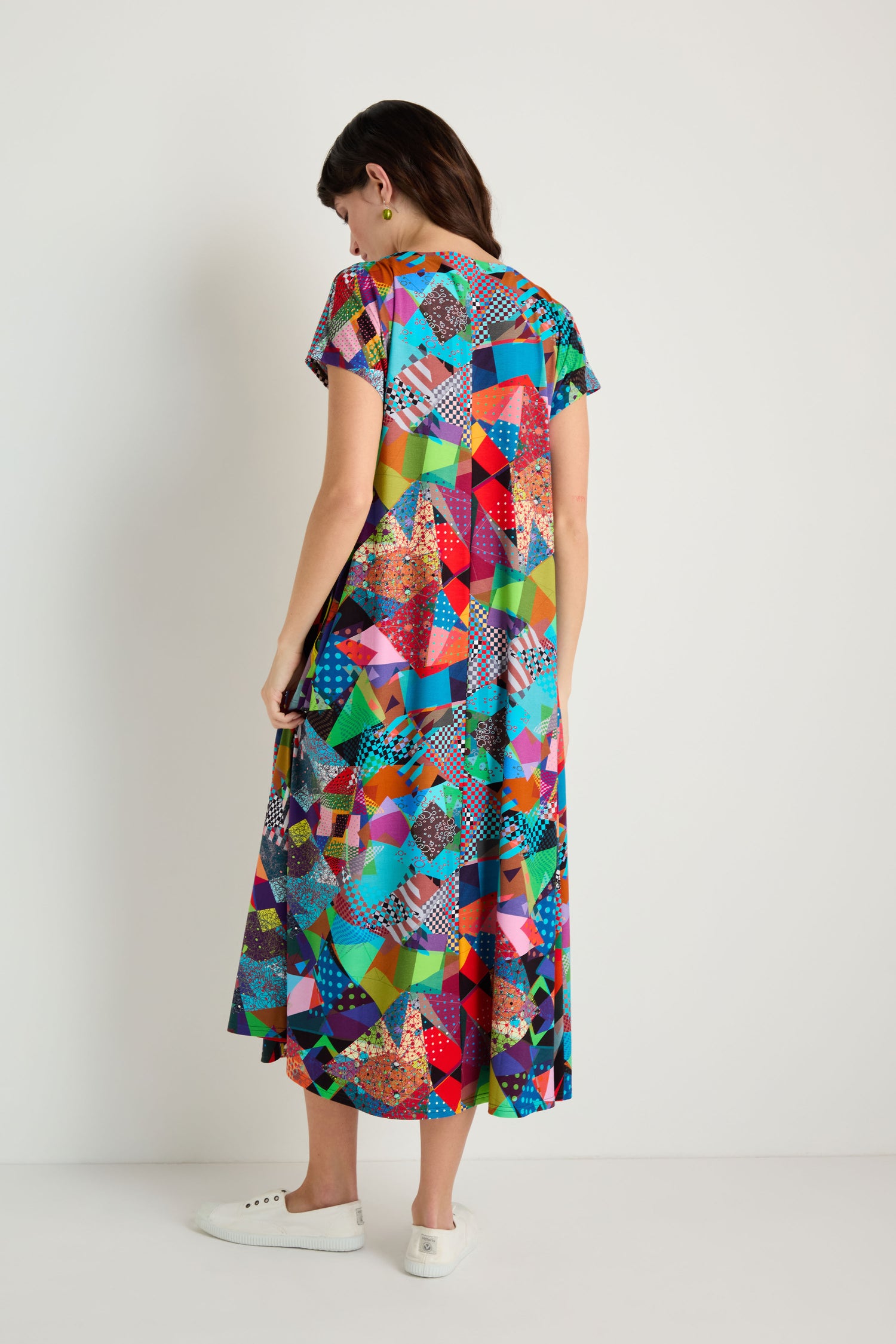 A woman stands facing away in a Quilt Print Flared Dress featuring a colorful geometric pattern, short sleeves, and paired with white sneakers against a plain background.