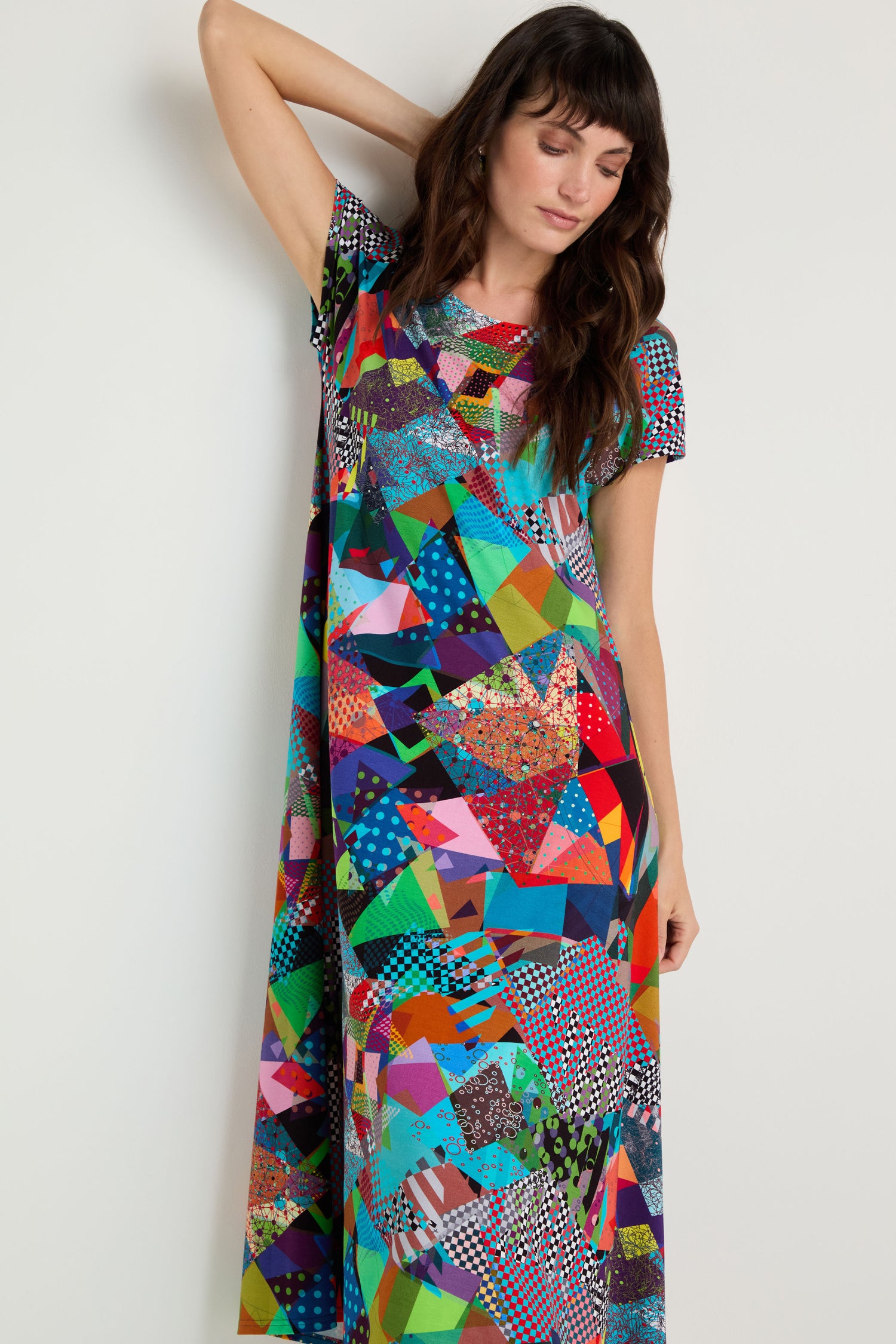 A woman poses against a white wall in the Quilt Print Flared Dress, featuring short sleeves and a vibrant quilt-inspired pattern. One arm is raised as she closes her eyes.