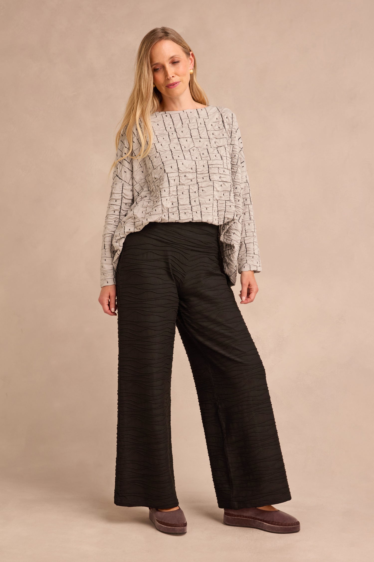 A woman with long blonde hair wears a loose patterned light gray top, Wave Textured Jersey Wide Leg Trousers in black, and mauve slip-on shoes, standing against a plain beige background.