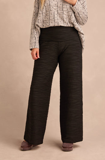 A person stands against a neutral background wearing a long-sleeve patterned top paired with Wave Textured Jersey Wide Leg Trousers, which feature a subtle raised wave pattern and a high-waisted silhouette.