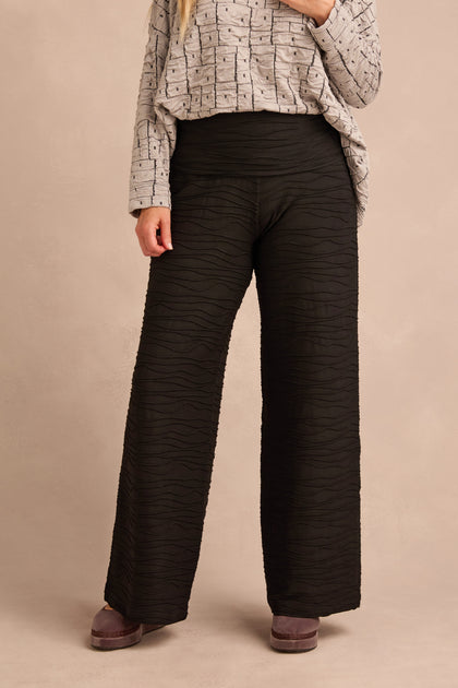 A person stands against a neutral background wearing a long-sleeve patterned top paired with Wave Textured Jersey Wide Leg Trousers, which feature a subtle raised wave pattern and a high-waisted silhouette.