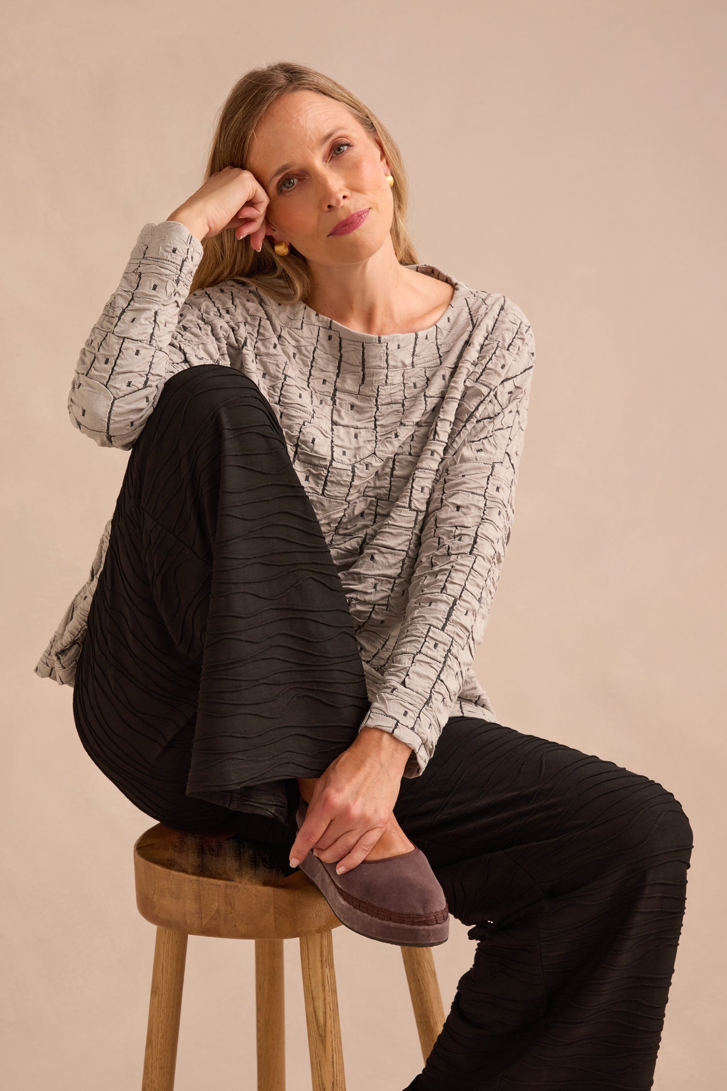 A woman sits on a wooden stool, resting her head on her hand and looking forward with a neutral expression, dressed in a gray patterned top and Wave Textured Jersey Wide Leg Trousers.