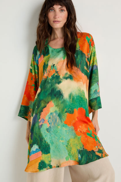 A woman stands against a plain white wall wearing the Japanese Garden Boxy Tunic, an oversized woven viscose top with an abstract green, orange, and blue pattern, paired with light beige pants.