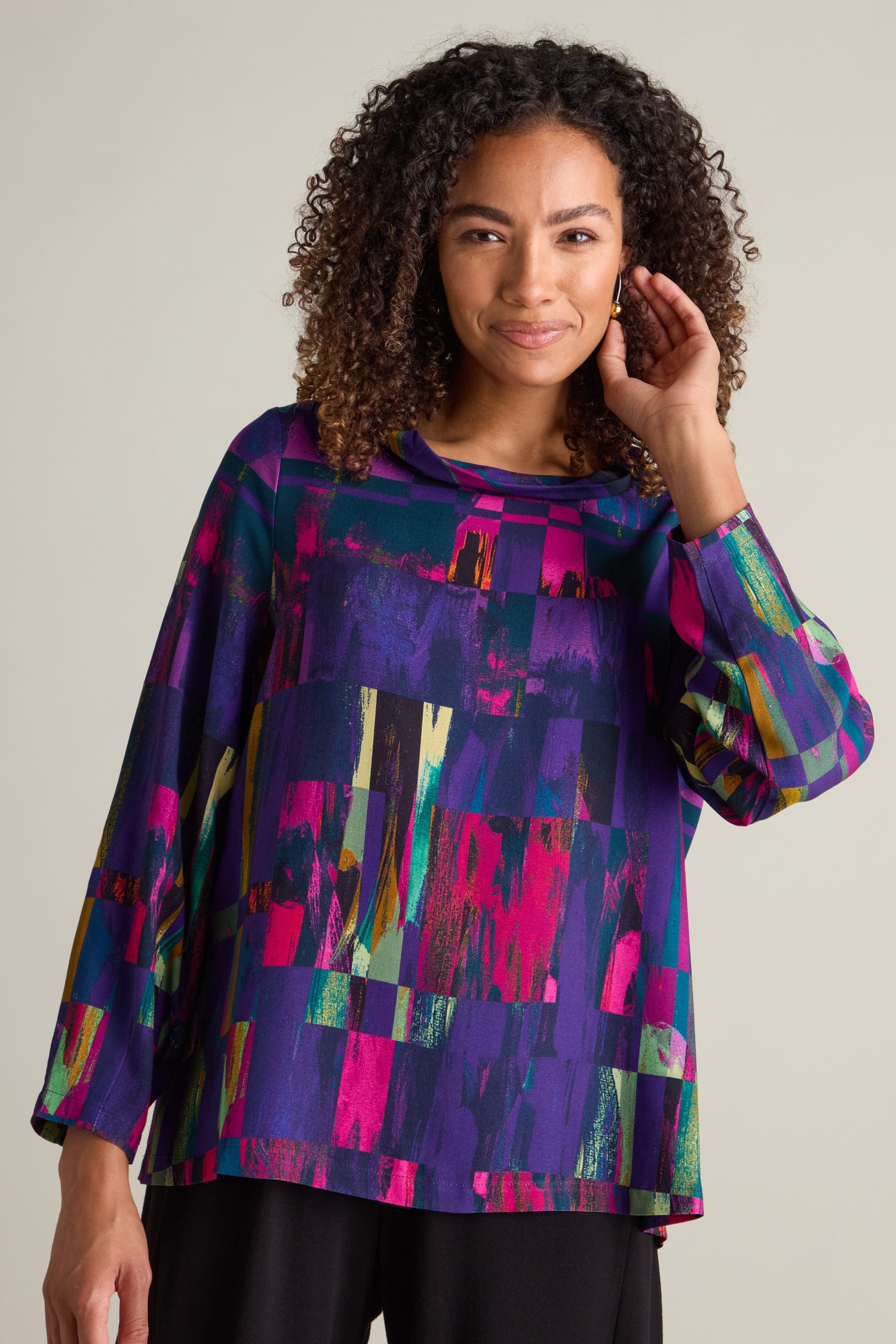 A person is smiling and touching their hair while wearing the Aliza Block Print Top, which features a vibrant, abstract pattern in rich jewel tones. The background is a plain, light-colored wall.