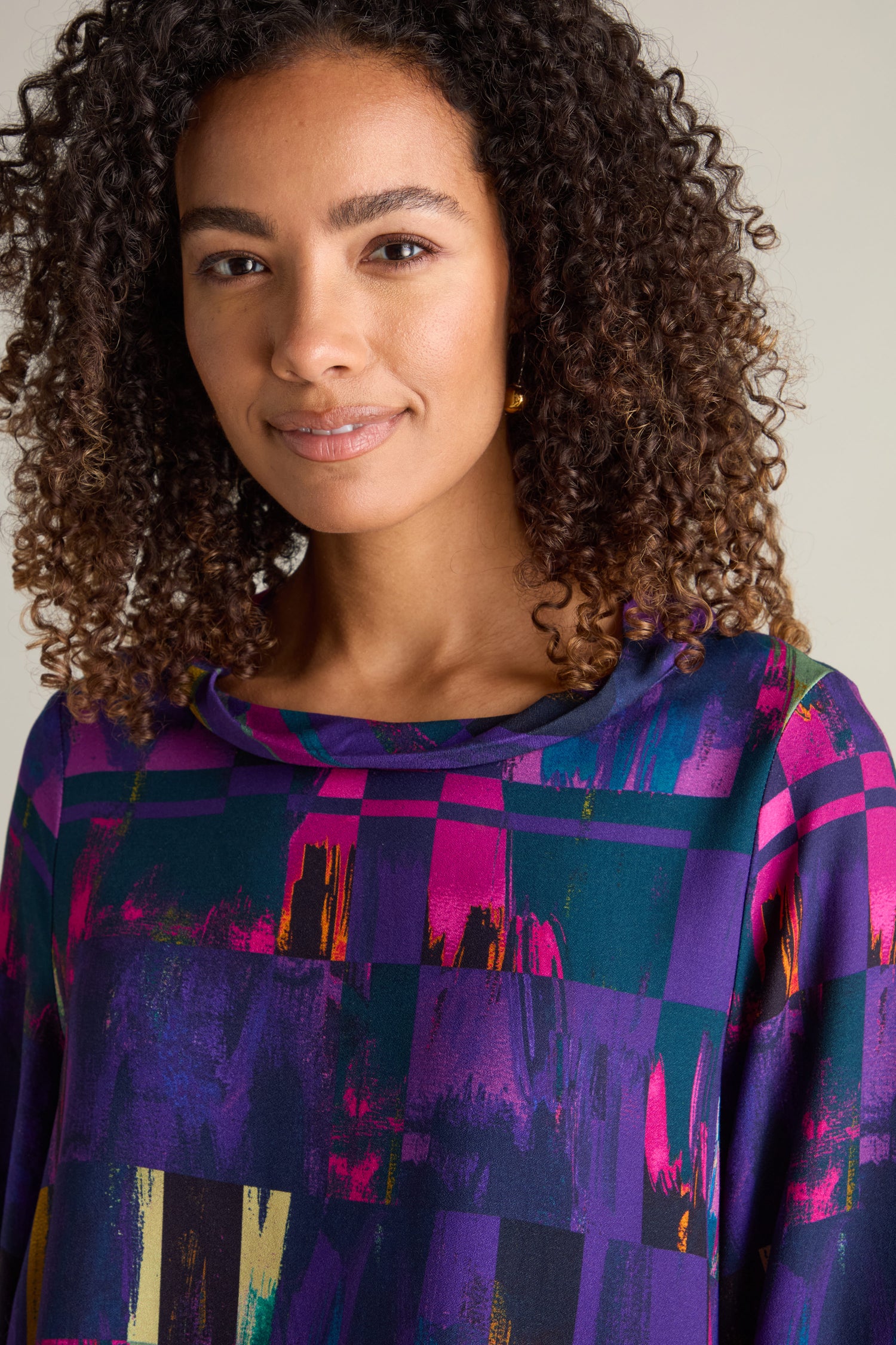 A woman with curly hair, wearing an Aliza Block Print Top adorned in vibrant jewel tones, gazes at the camera with a subtle smile.