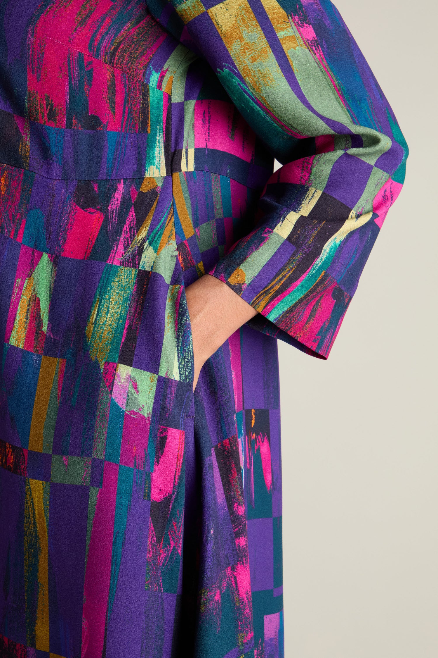 Close-up of a person's arm and hand in a pocket of the exquisite Malina Block Print Dress, showcasing its brightly colored, abstract-patterned garment adorned in vibrant shades of purple, green, yellow, and pink.