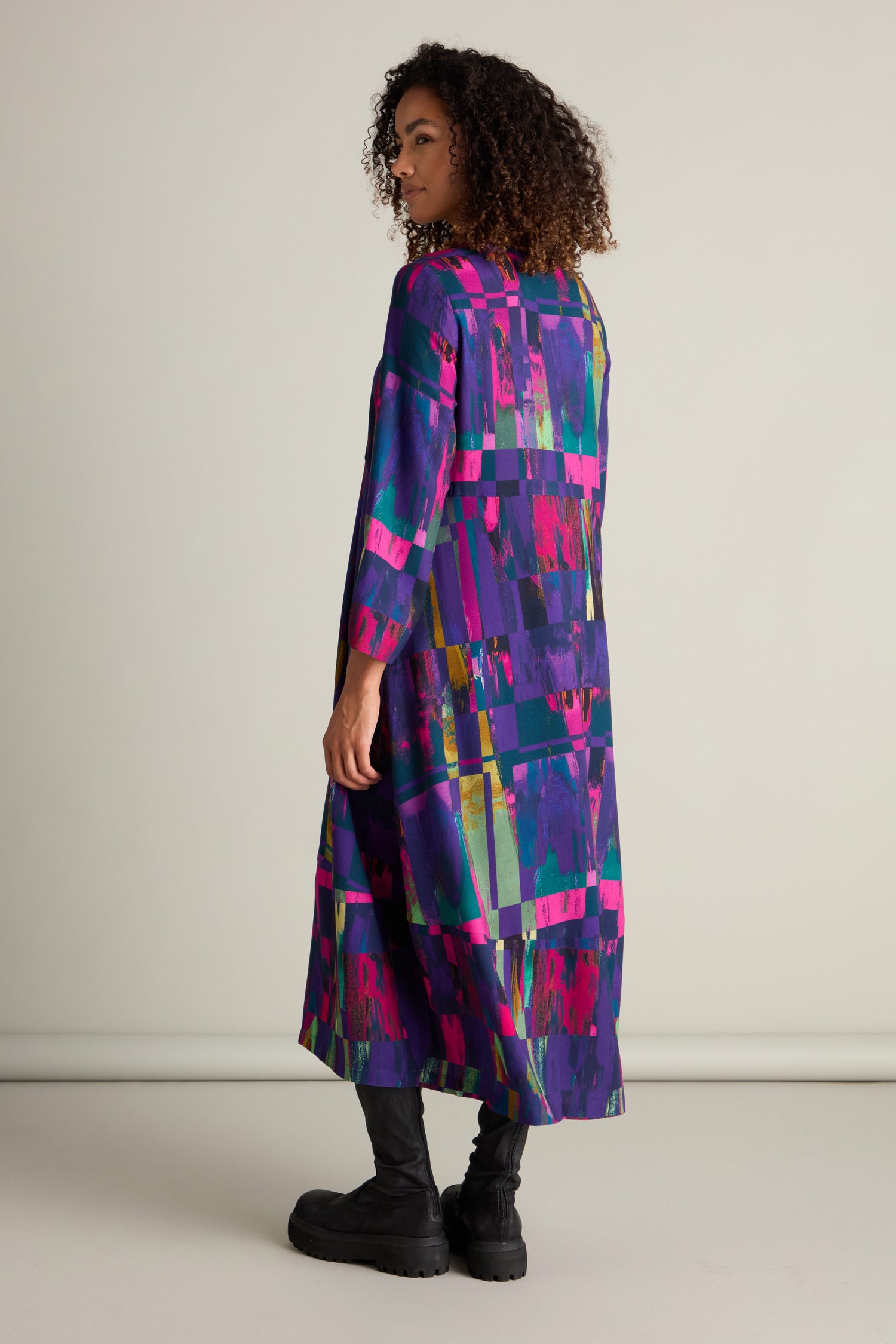 Person standing and facing sideways, wearing the stunning Malina Block Print Dress adorned with vibrant abstract patterns and paired with black boots, highlighting its exquisite craftsmanship against a plain background.