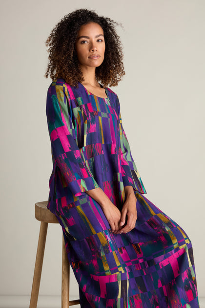 A woman with curly hair is seated on a wooden stool, wearing the long and colorful Malina Block Print Dress, adorned with abstract patterns. She faces the camera with a neutral expression, perfectly showcasing the exquisite craftsmanship of her attire.