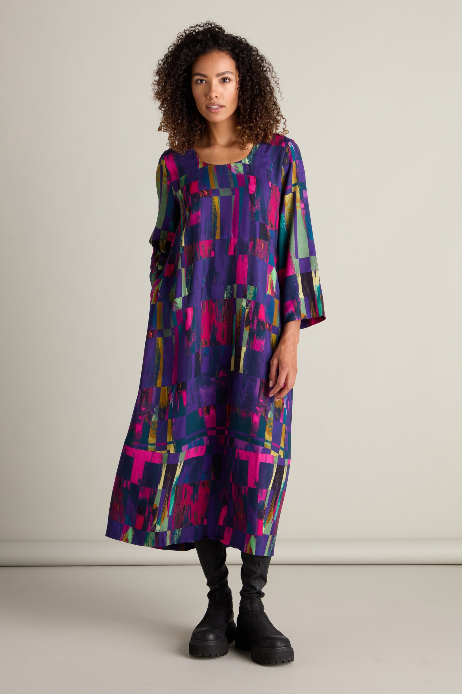 A person with curly hair wears the vibrant Malina Block Print Dress paired with black boots, highlighting its exquisite craftsmanship against a plain backdrop.