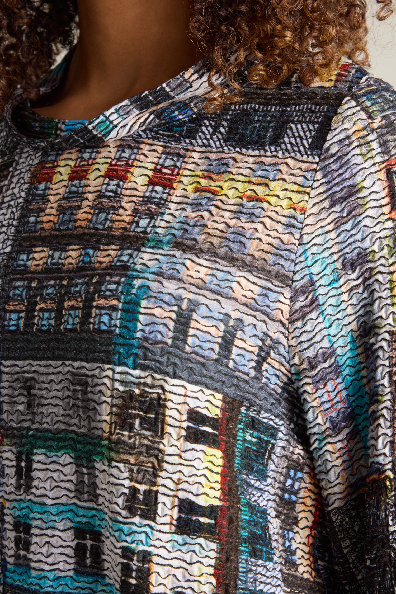 Close-up of a person wearing the Aliza Facade Print Jersey Top, showcasing its vibrant abstract patterns with wavy lines and geometric shapes. The person's curly hair peeks into the frame. This versatile top adds architectural intrigue to everyday fashion, making it a standout piece in any wardrobe.