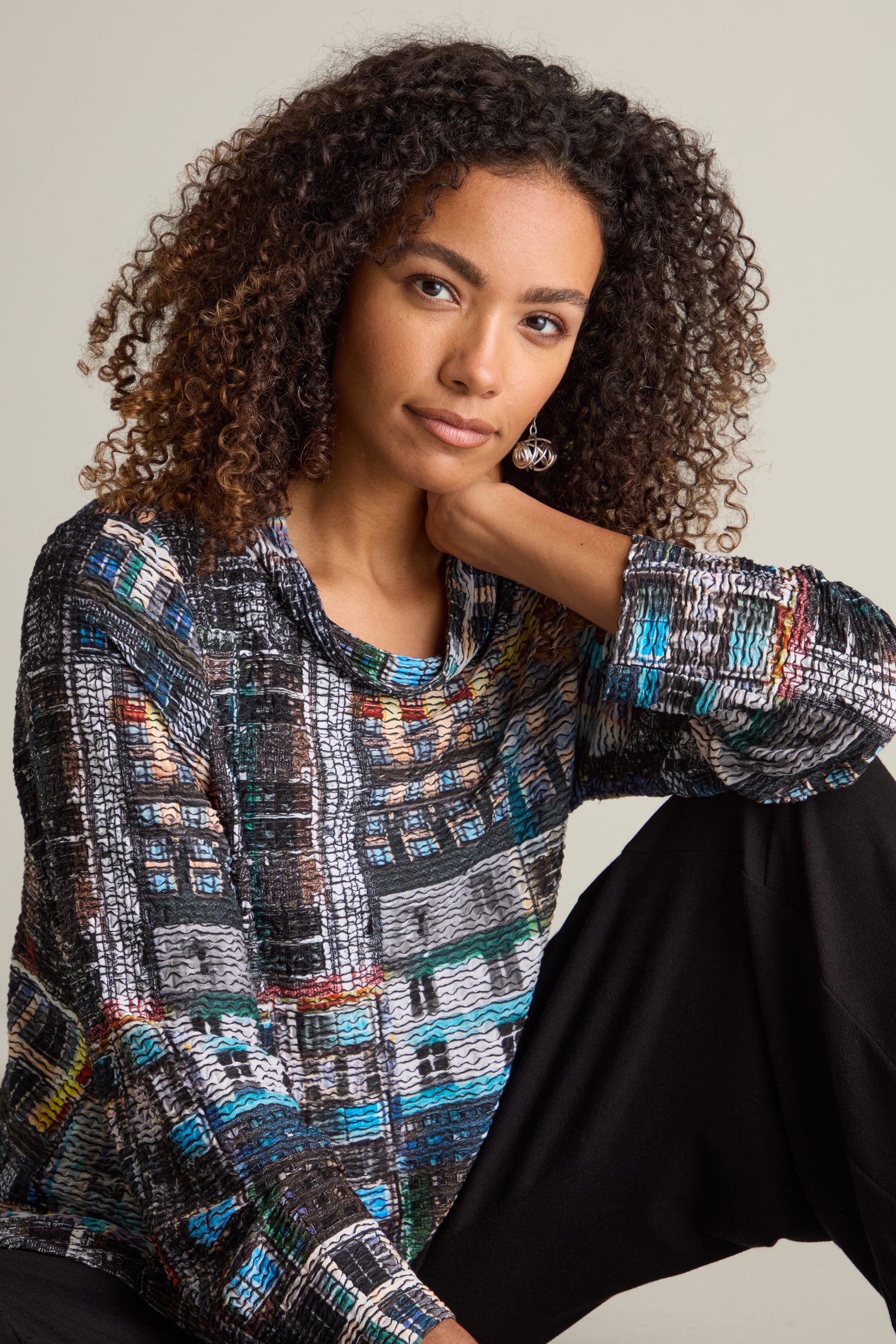 A woman with curly hair, adorned in an Aliza Facade Print Jersey Top and black pants, rests her head on her hand. The architectural intrigue of the top’s versatile design enhances her look.