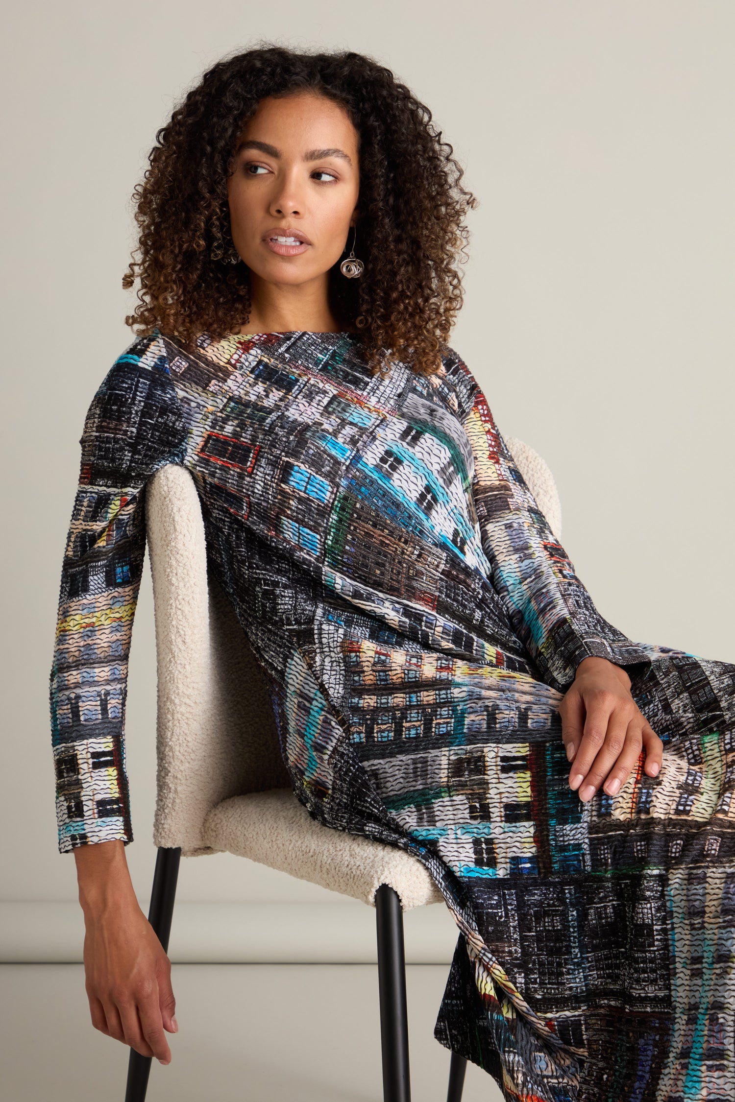 A woman with curly hair, dressed in the Ramona Facade Print Jersey Dress featuring long sleeves and a voluminous hem adorned with a multi-colored abstract print, sits on a white chair against a beige background.