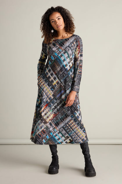 A woman with curly hair is wearing the Ramona Facade Print Jersey Dress with a long, boat neckline, paired with black boots, standing against a plain background.