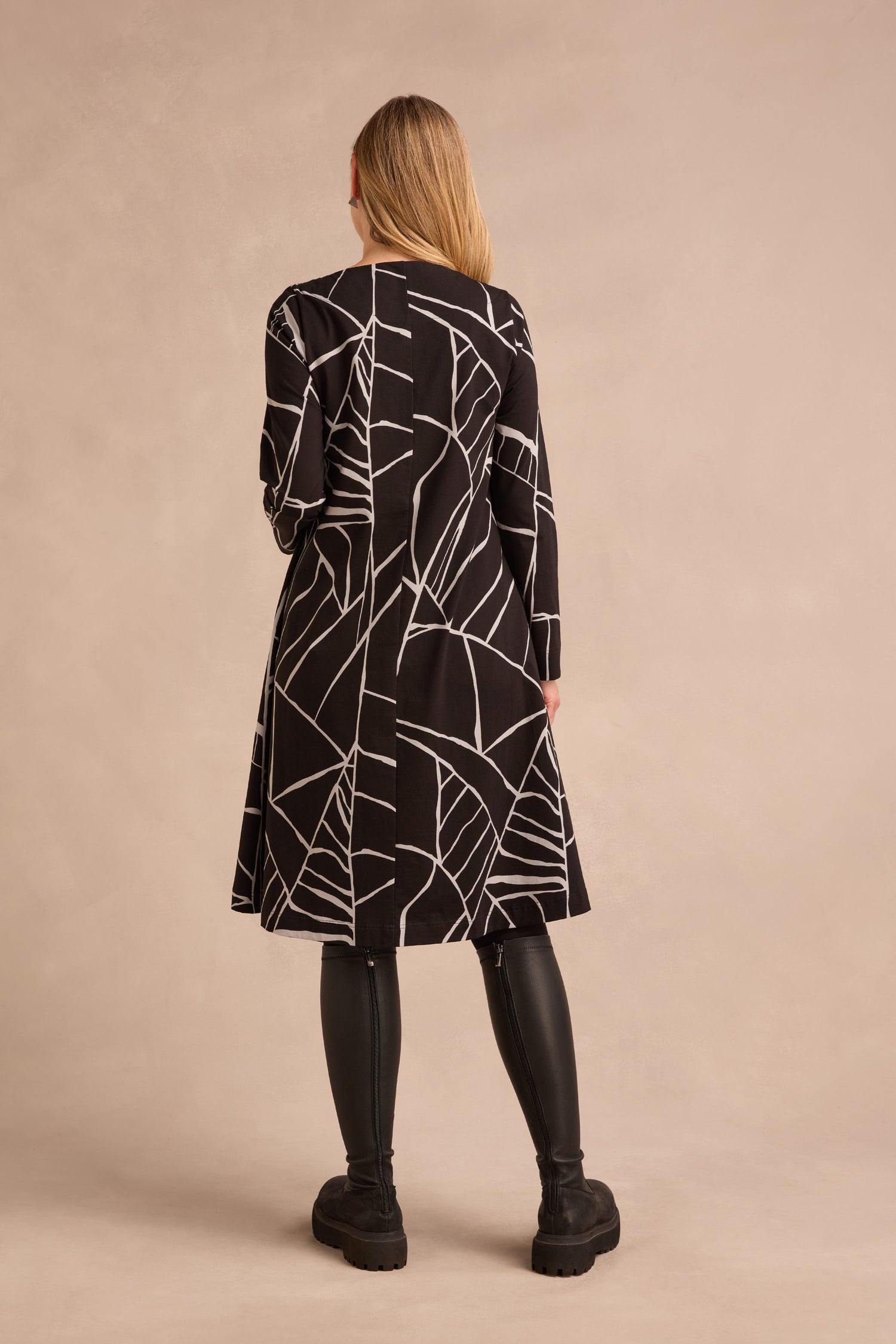 A person stands with their back to the camera, wearing the Linear Print Jersey Flared Dress in black with white geometric lines, paired with black leggings and black boots, set against a plain beige background.
