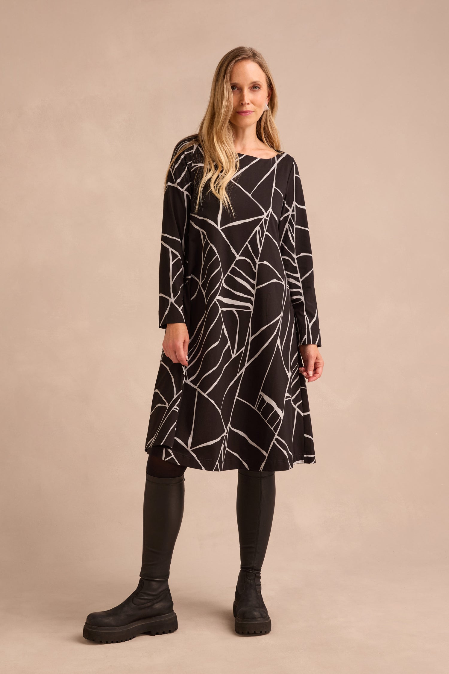 A woman stands against a neutral background wearing the Linear Print Jersey Flared Dress, styled with black leggings and boots.