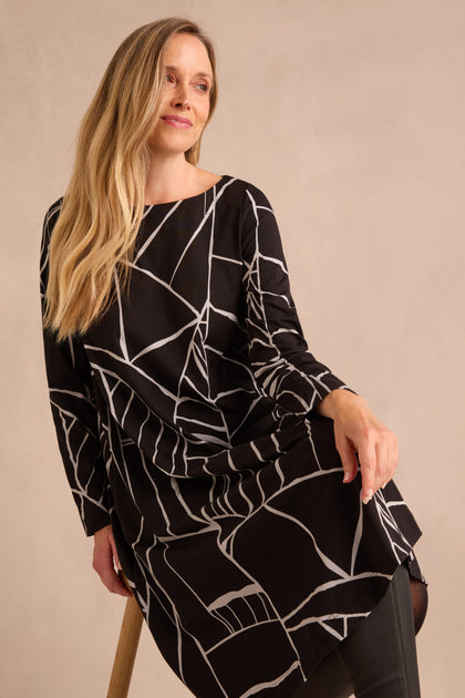 A woman with long blonde hair wears the Linear Print Jersey Flared Dress, featuring black fabric with white abstract lines, as she sits on a wooden stool against a plain beige background.