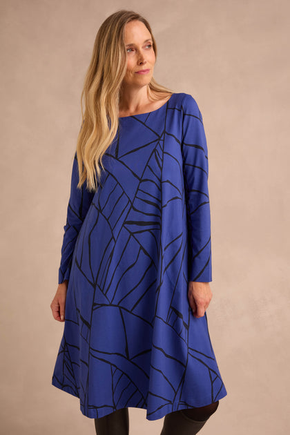 A woman wearing the Linear Print Jersey Flared Dress Aino, a long-sleeved, knee-length blue dress featuring a black abstract line pattern, stands against a plain beige background.