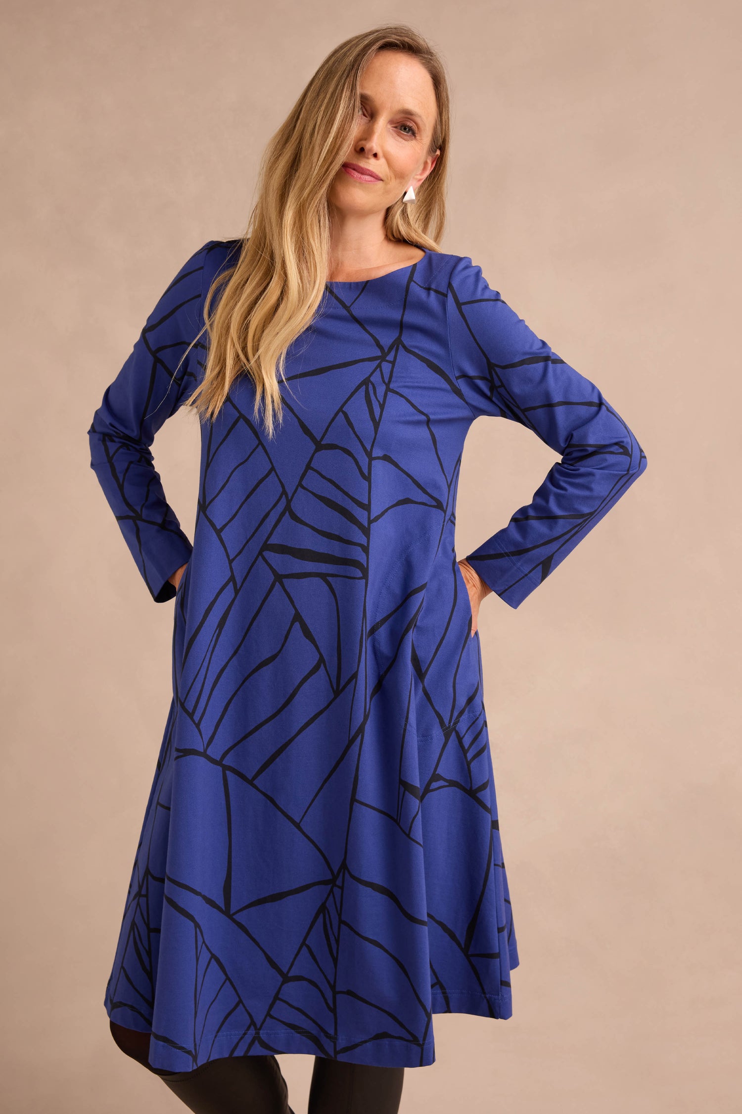 A woman stands with her hands on her hips against a neutral background, wearing the Linear Print Jersey Flared Dress Aino—a long-sleeved, knee-length dress in blue featuring a black abstract line pattern.