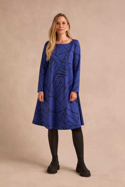 A woman stands against a neutral beige background wearing the Linear Print Jersey Flared Dress Aino, featuring black geometric lines, styled with black leggings and black boots.