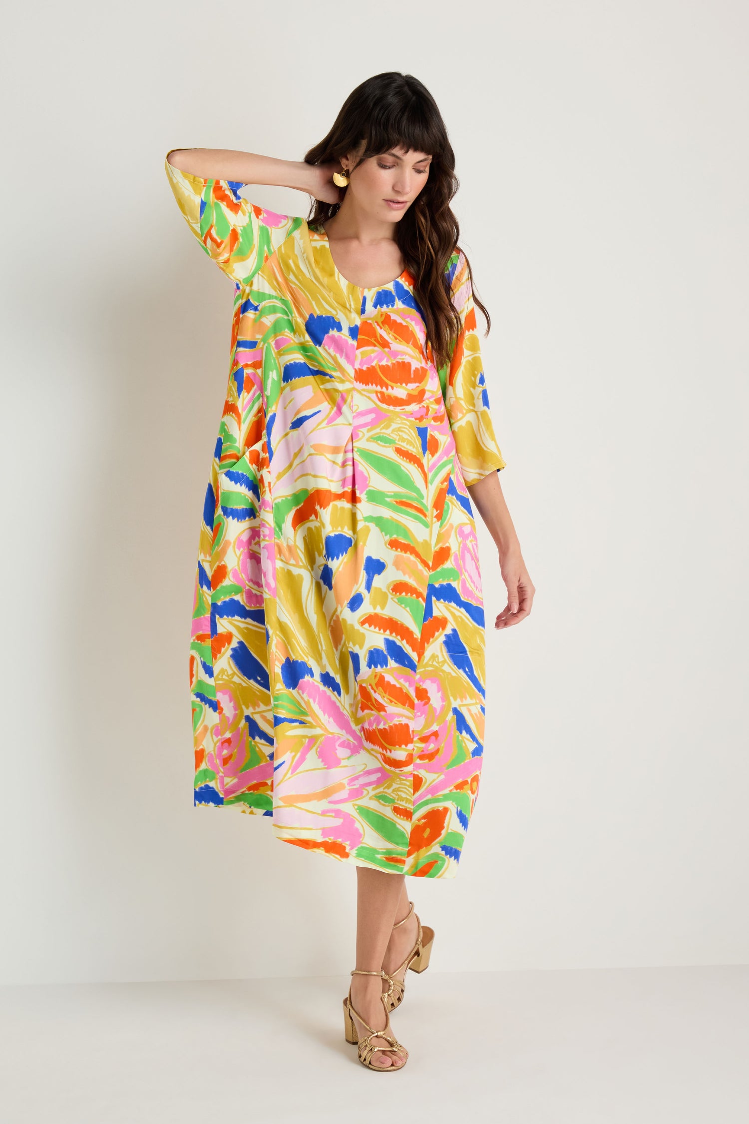 A woman models the Abstract Floral Pleated Dress with gold heels, posing against a plain white background and resting one hand on her head.