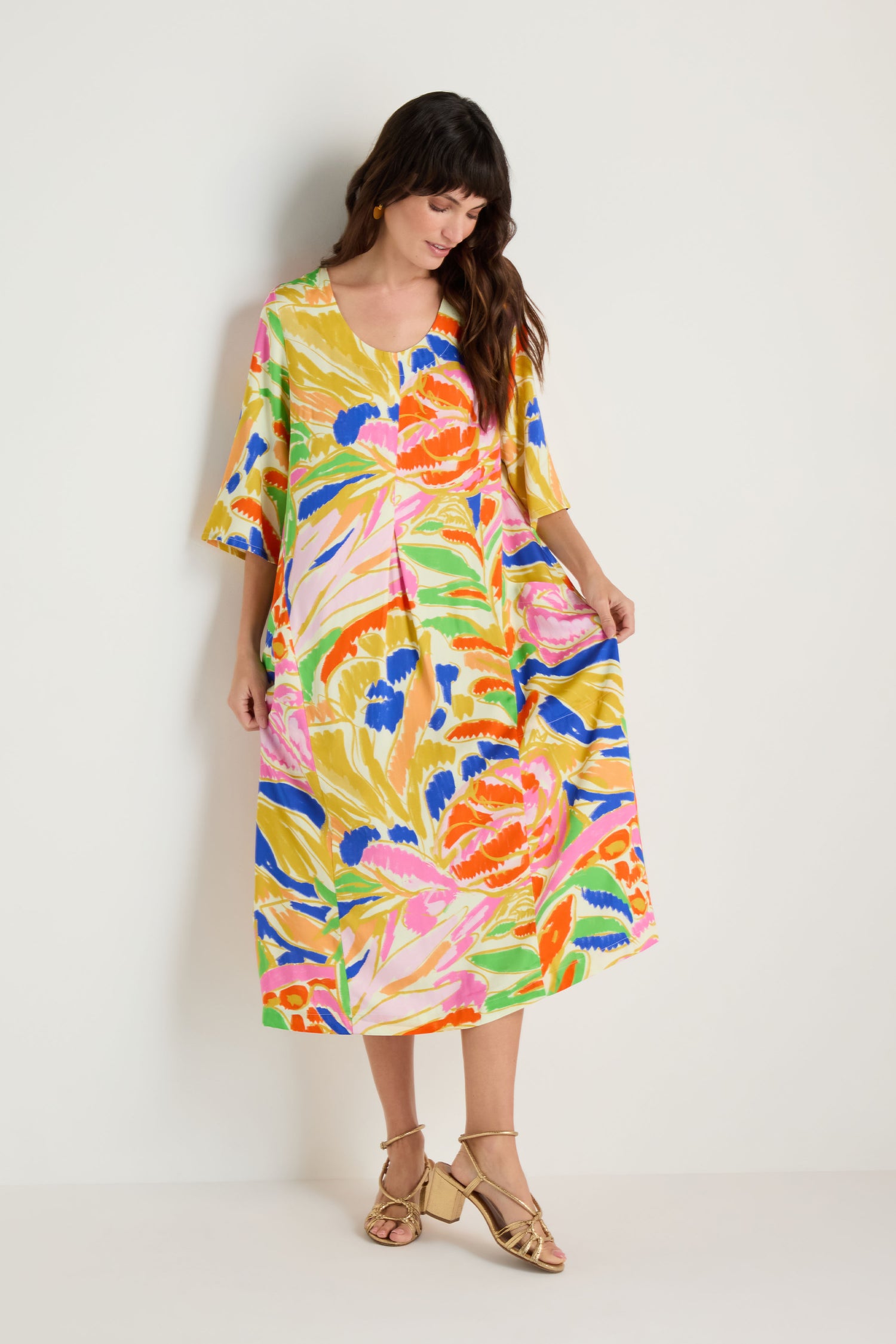 A woman stands by a plain wall, looking down and holding the sides of her Abstract Floral Pleated Dress in midi length with short sleeves, paired with gold sandals.