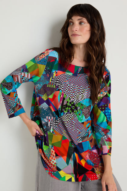 A woman wears the Quilt Print Flared Top, featuring long sleeves and a flared shape, paired with grey pants. She stands against a plain light wall and gazes to the side.