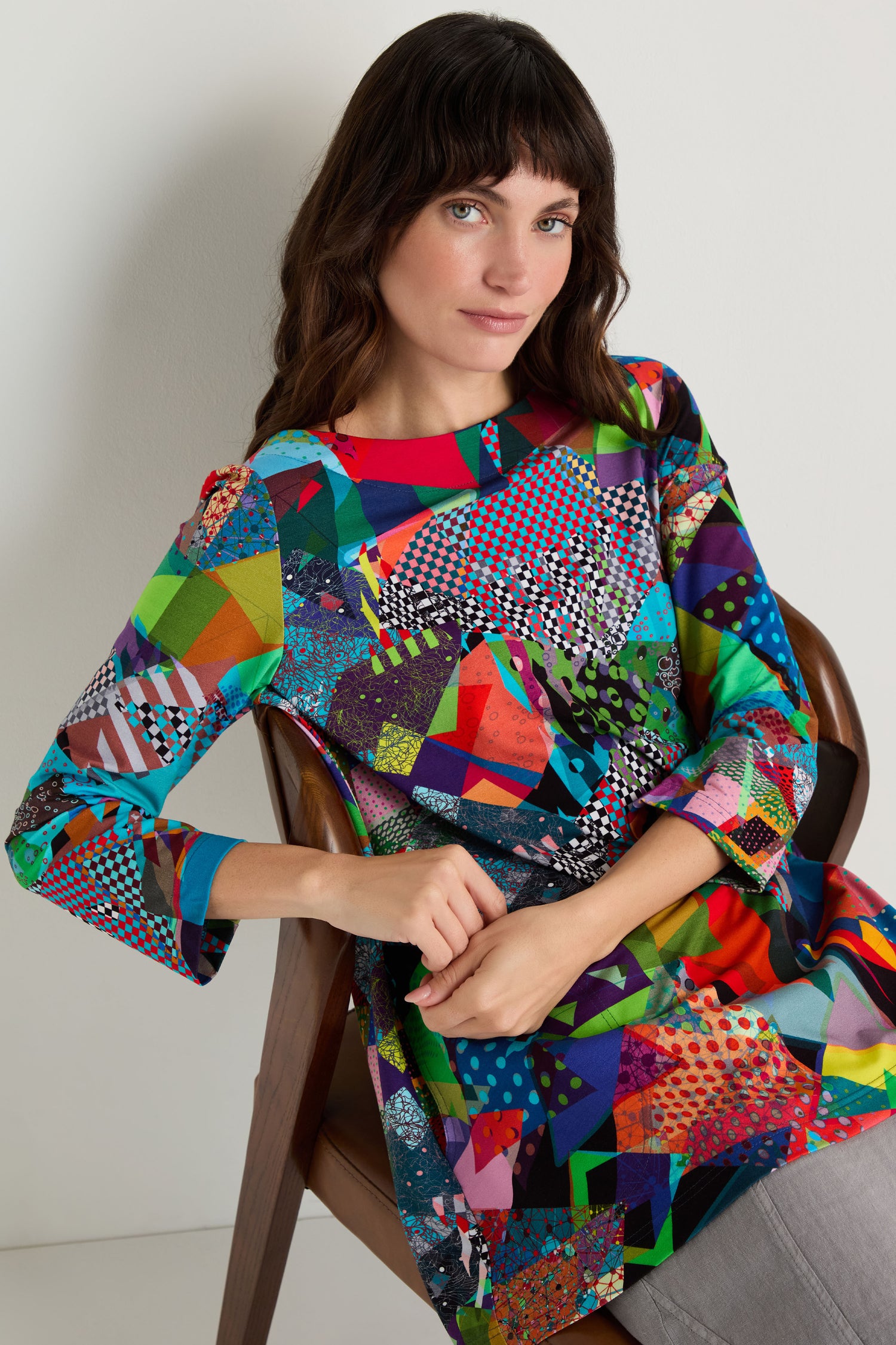 A woman with straight brown hair sits on a wooden chair in front of a plain white wall, wearing the Quilt Print Flared Top and gray pants.