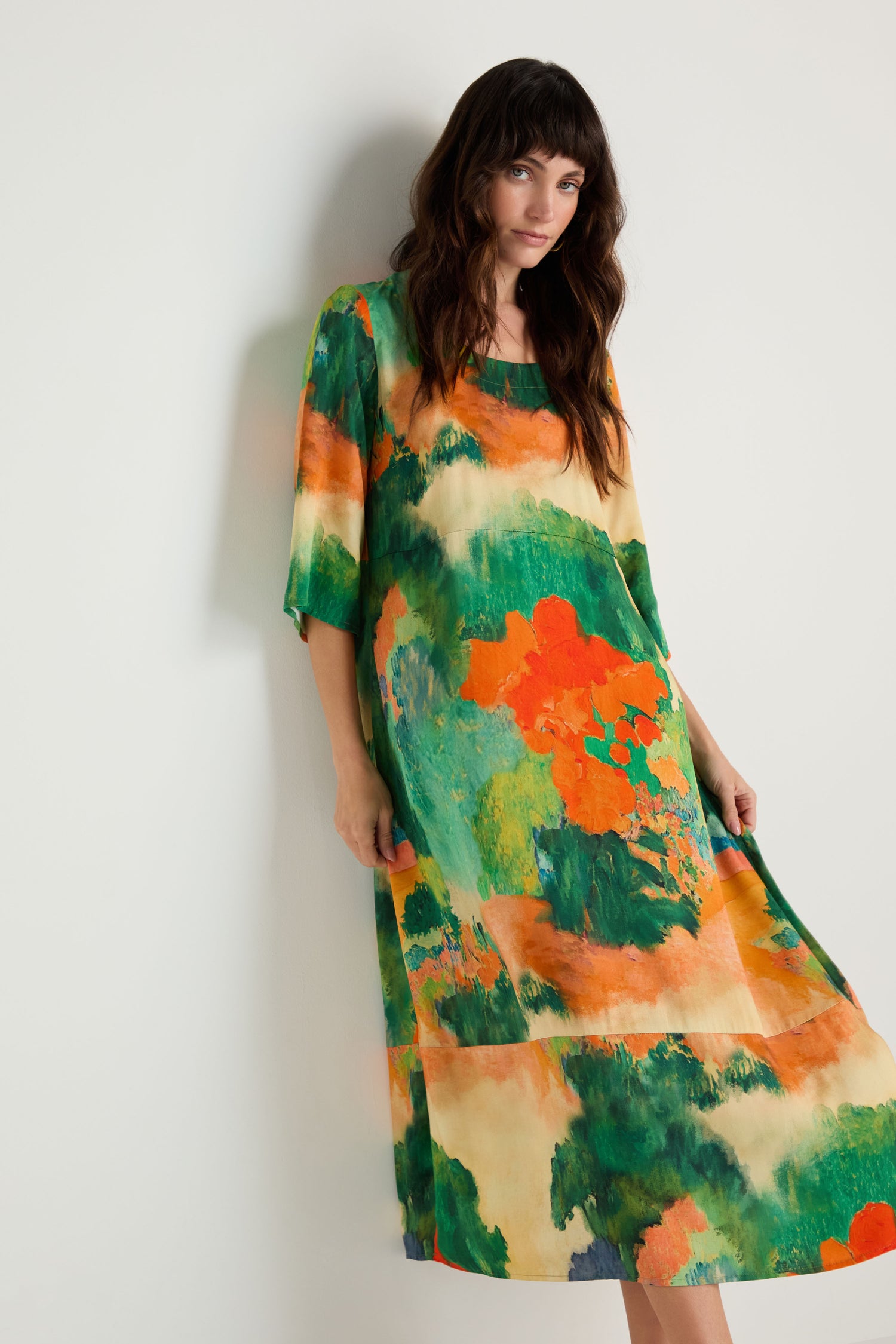 A woman models the Japanese Garden Flared Dress, a loose-fitting, mid-length piece with a green, orange, and beige abstract pattern, standing against a plain white wall.
