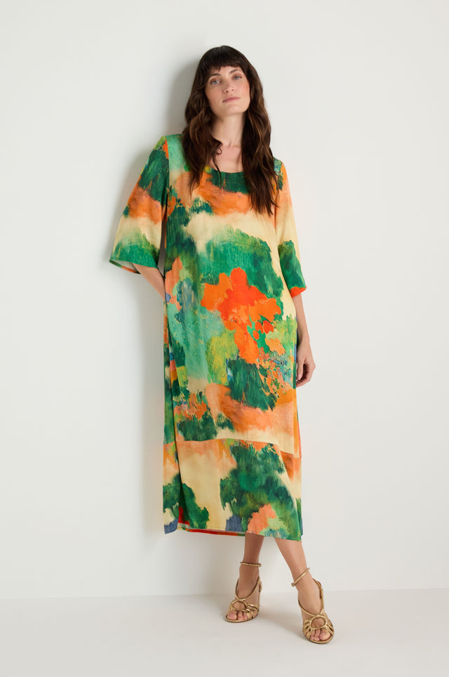 A woman stands against a plain wall wearing the Japanese Garden Flared Dress in green, orange, and beige abstract patterns with beige strappy sandals. The ankle-length dress is made from soft woven viscose.
